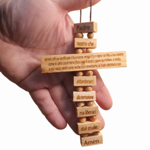 Una mano sorregge una croce artigianale fatta di piccoli blocchi rettangolari e grani tondi in legno d'ulivo chiaro. Su ogni blocco è incisa una parte della preghiera del "Padre Nostro" in lingua italiana. La croce pende da un cordino marrone ed è caratterizzata dalle venature naturali tipiche del legno della Terra Santa.